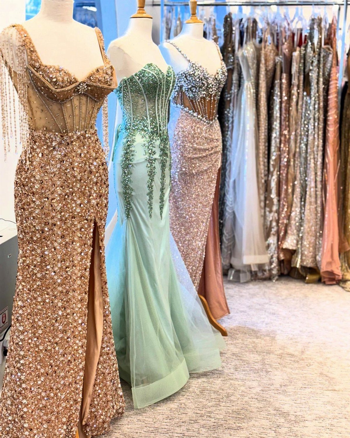 Three sequined mannequins wearing long, elegant prom dresses in a brightly lit clothing boutique.