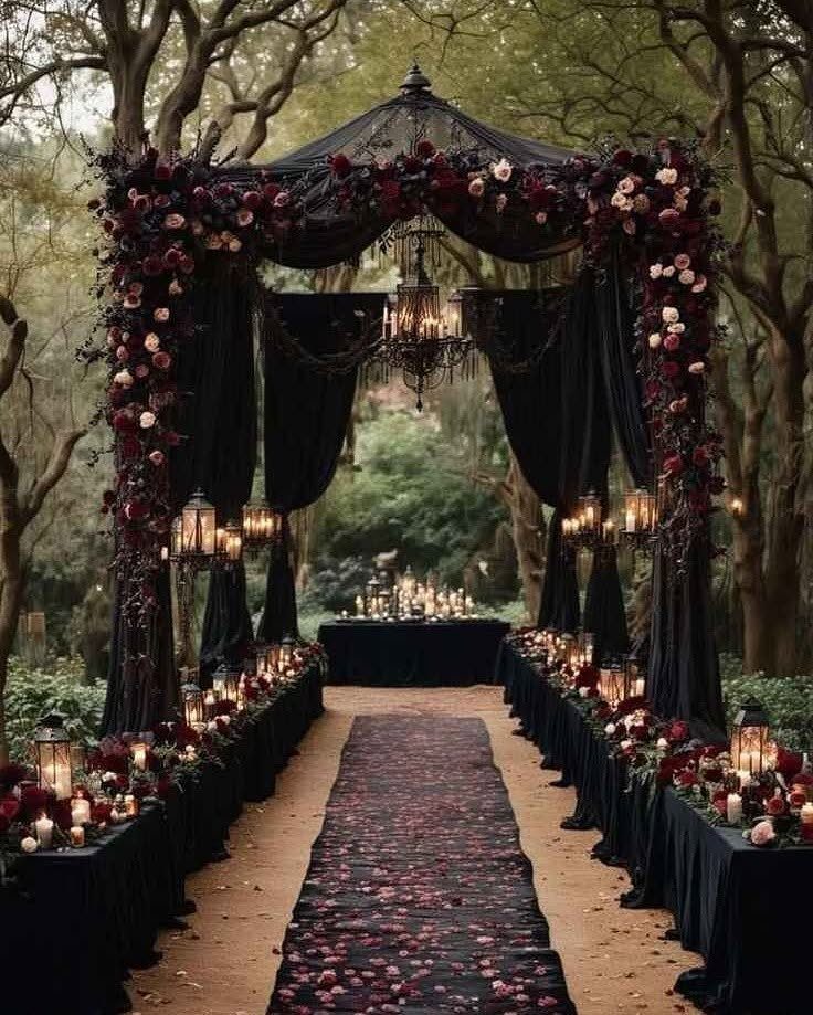 A black floral arch draped in red roses and black fabric over a candlelit aisle.