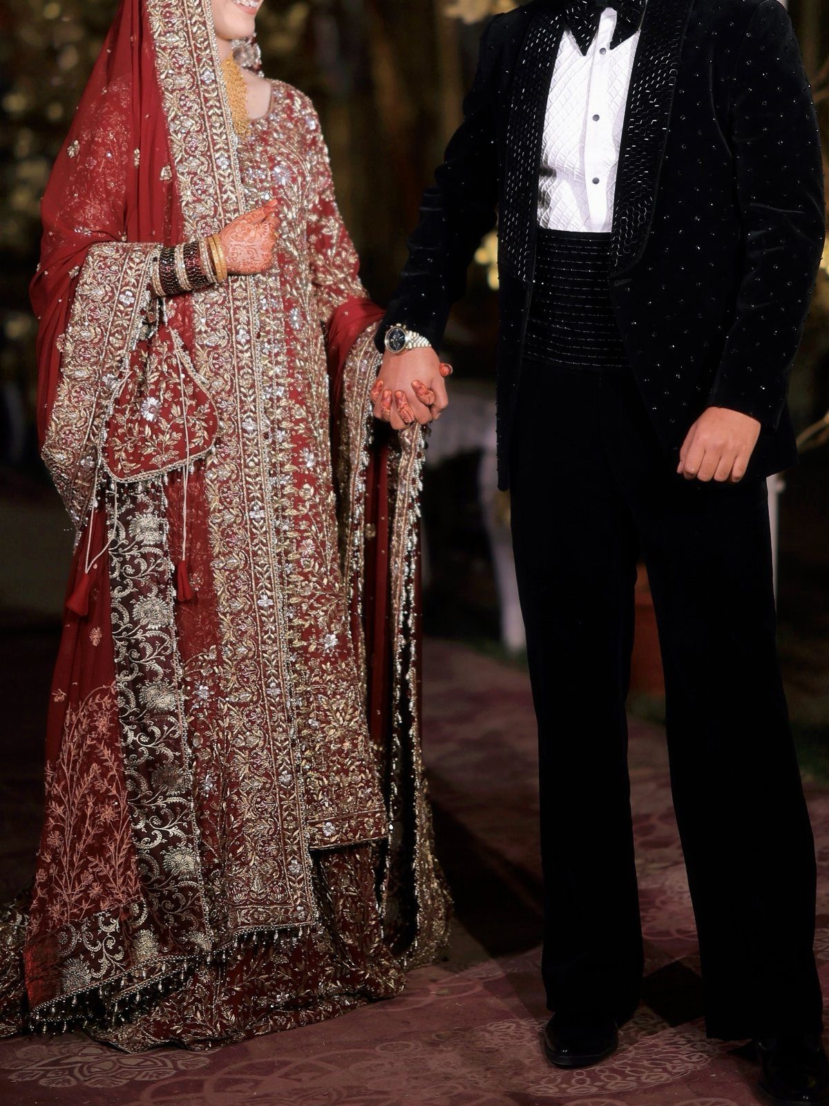 A bride in a red embroidered lehenga and a groom in a black tuxedo holding hands.