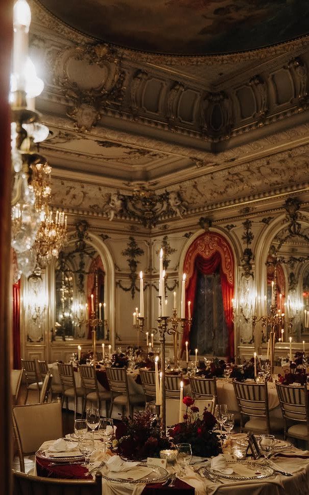 Ornate ballroom featuring red velvet curtains in alcoves and tall gold candelabras on dining tables.