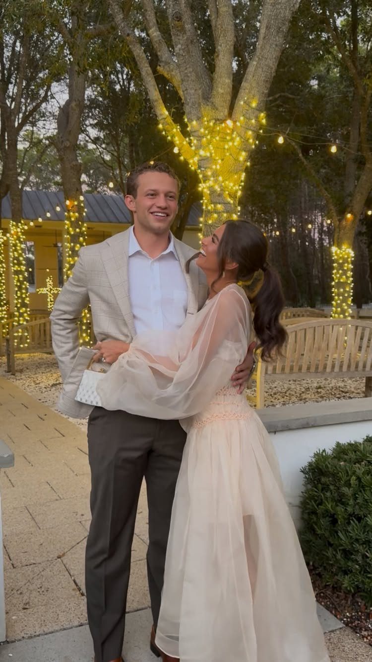 A woman in a peach balloon sleeve dress smiling at a man in a grey blazer outdoors.
