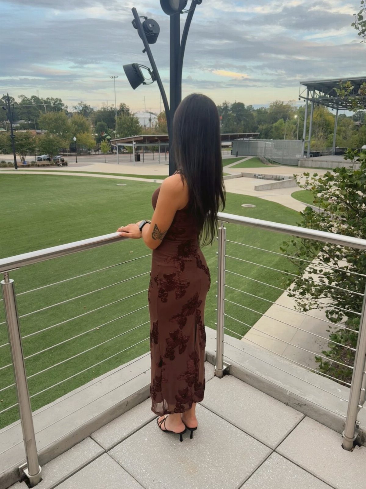 Woman in a brown floral maxi dress and black heels standing on a balcony overlooking a park.