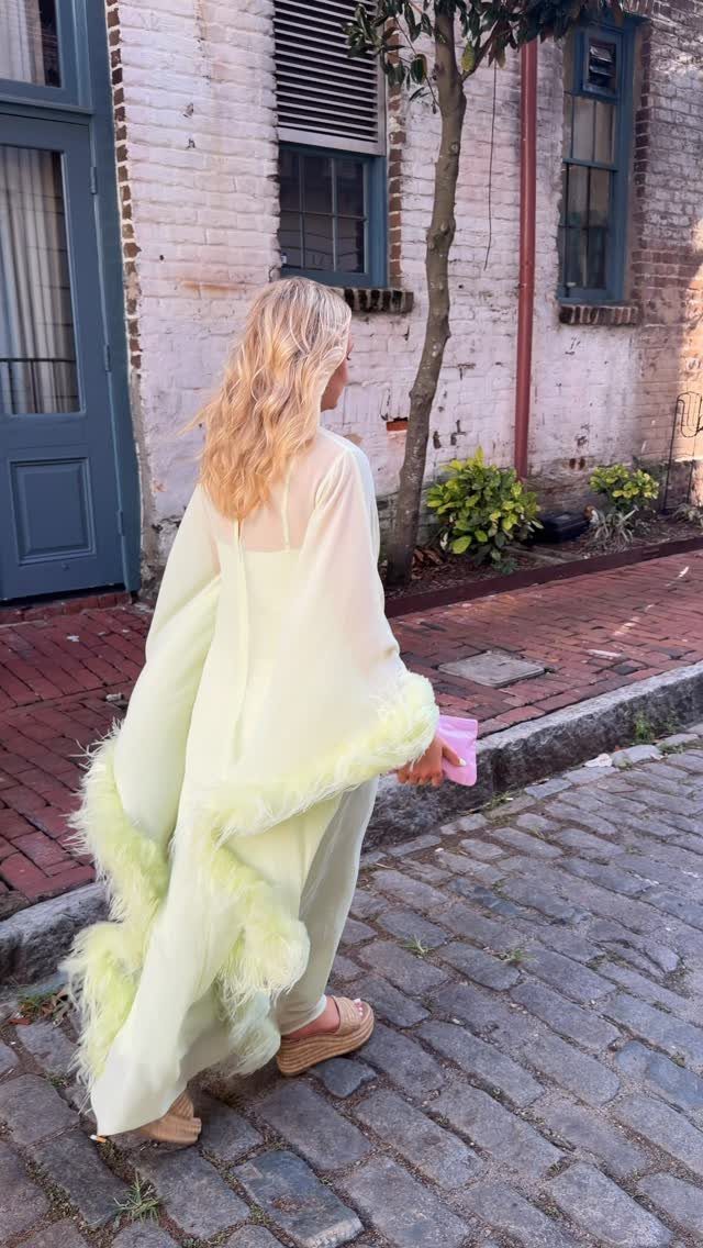 Blonde woman walking on a cobblestone street wearing a lime green jumpsuit with feather trim.