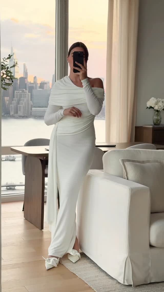 Woman in a long white off-the-shoulder draped dress posing in front of a city skyline view.