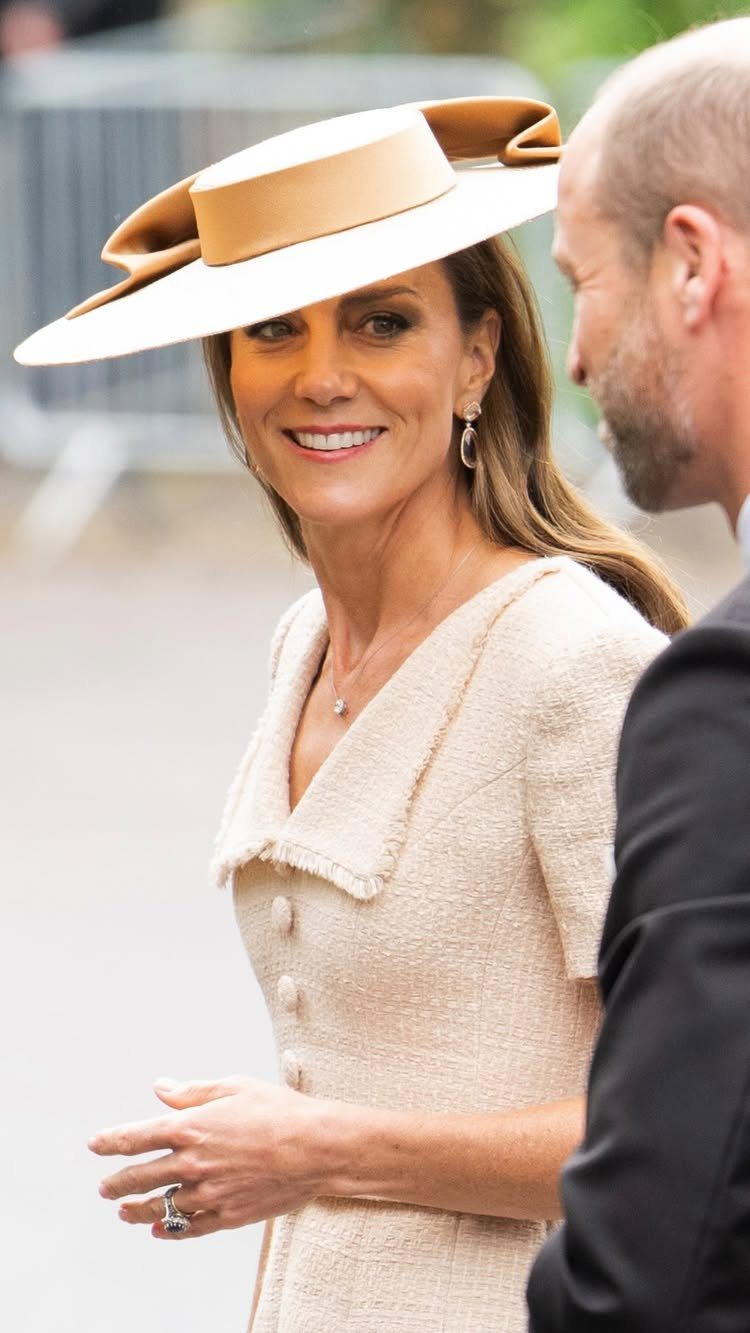 A woman in a cream dress and large tan hat smiling while talking to a man.
