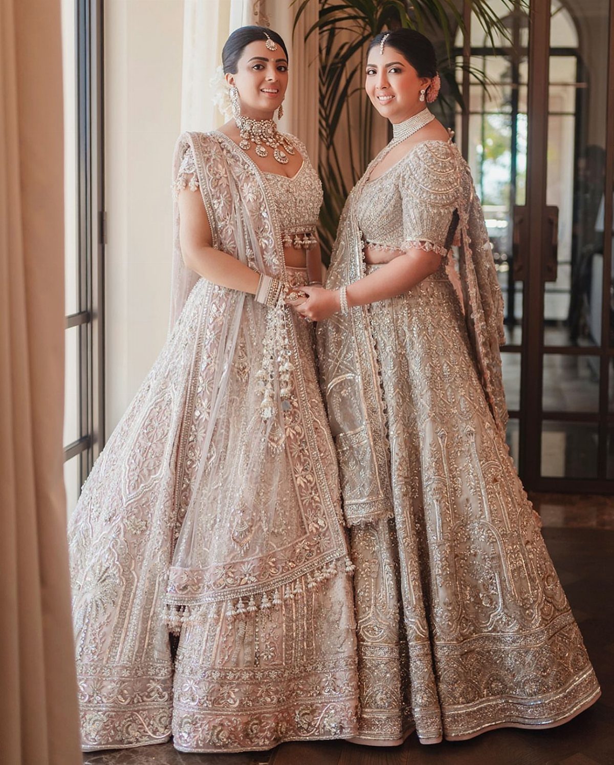 Two women wearing heavily embroidered silver traditional Indian lehengas and ornate jewelry while posing together.