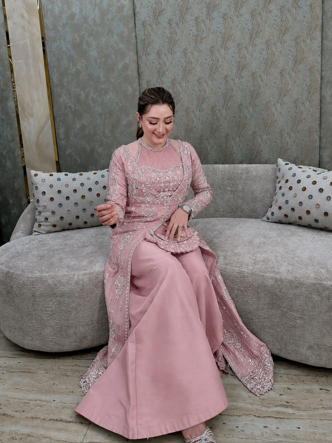 Woman in an embellished pink tunic and wide-leg trousers sitting on a grey sofa.
