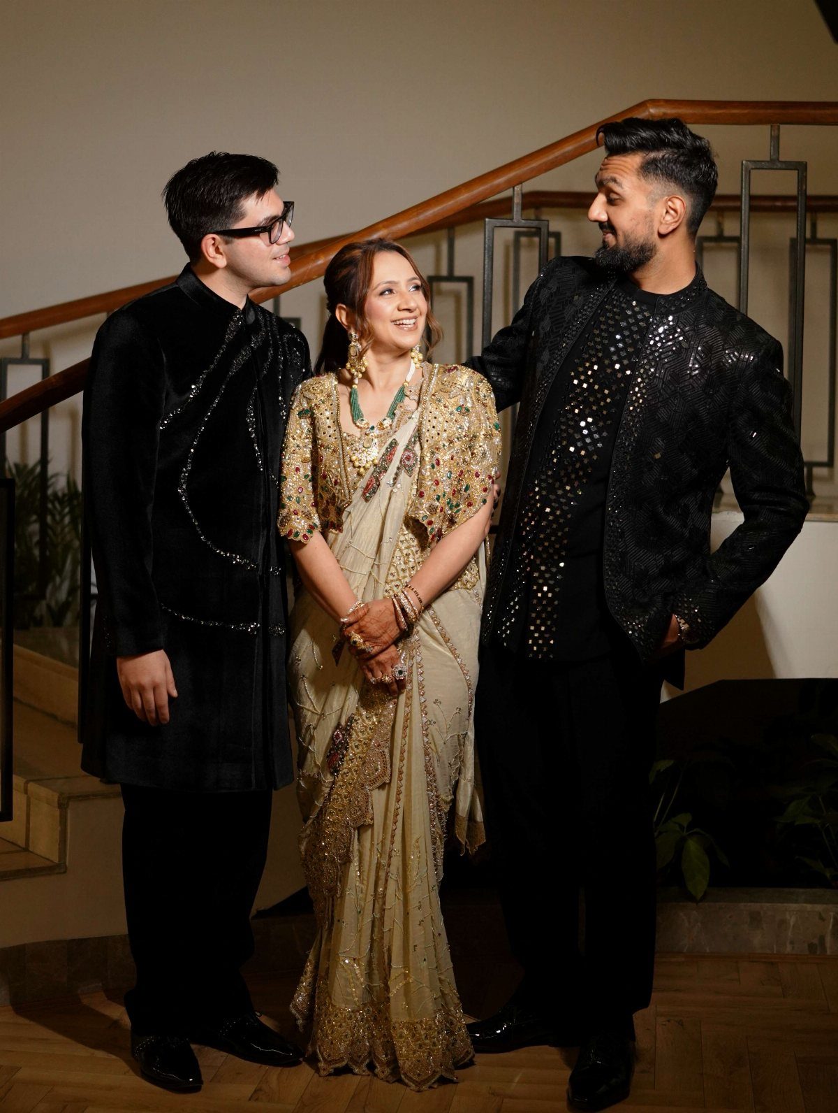 Three people dressed in elegant formal South Asian attire posing together indoors near a staircase.