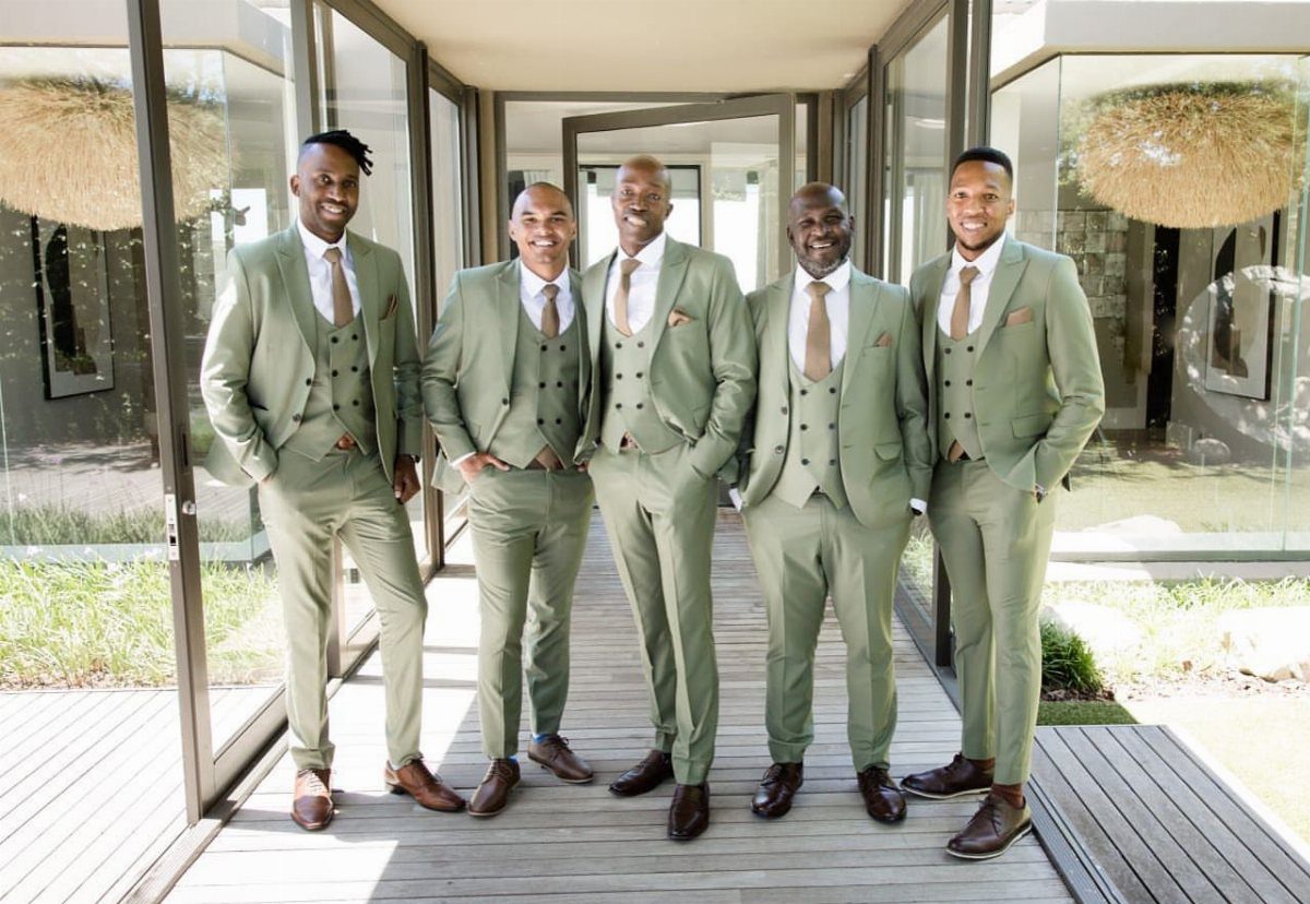 Five men posing in matching sage green three-piece suits on a wooden walkway.