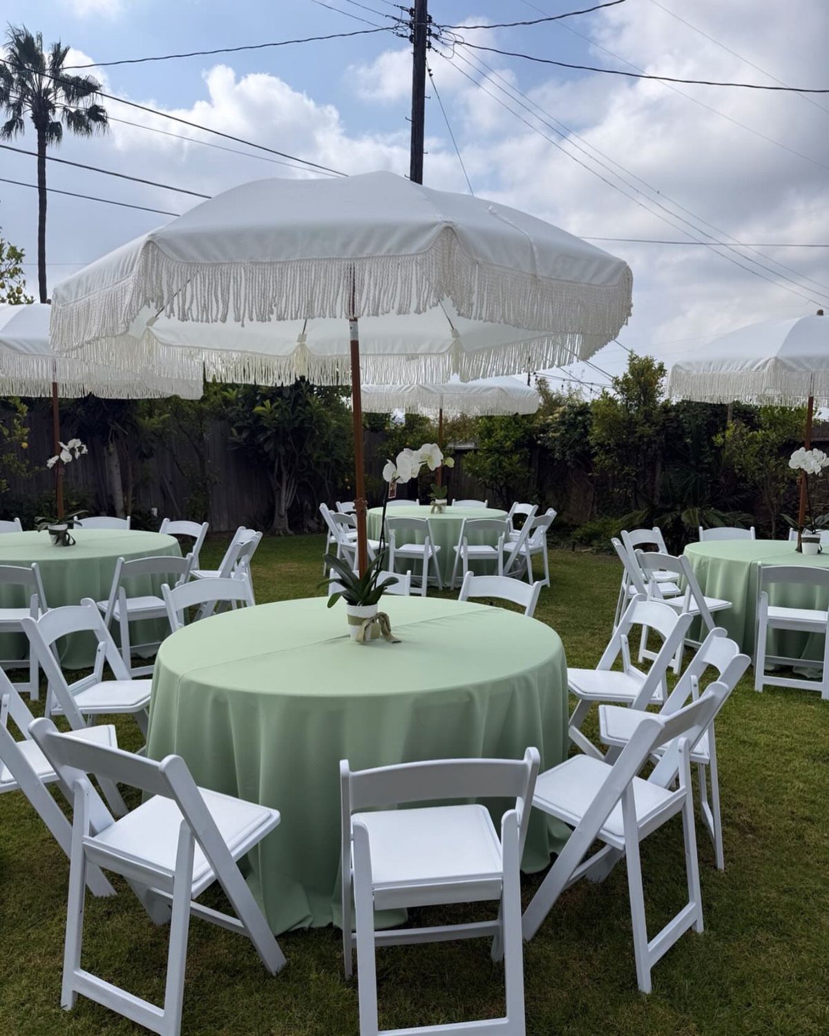 White fringed umbrellas shade round tables with sage green tablecloths and white chairs outdoors.