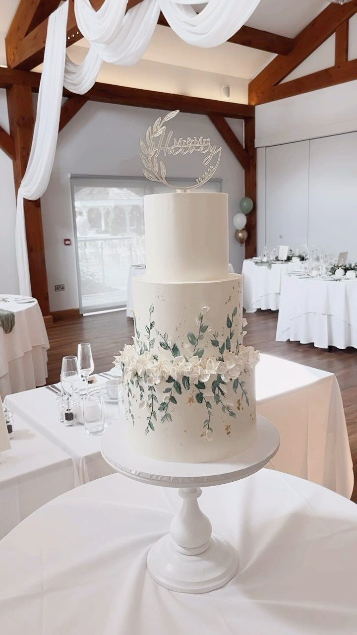 A white three-tiered wedding cake decorated with sage green leaves and white floral accents.