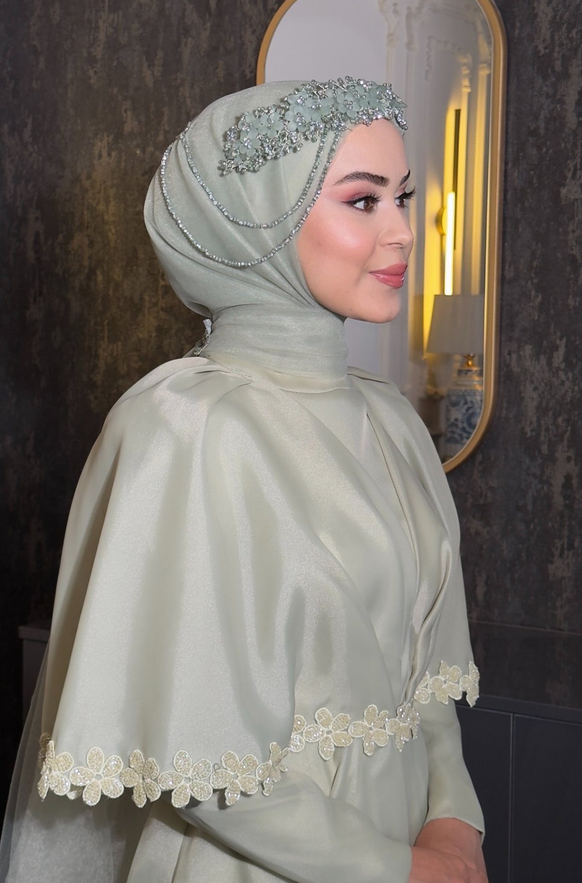 A woman in a sage green hijab and satin cape wearing a beaded floral headpiece.