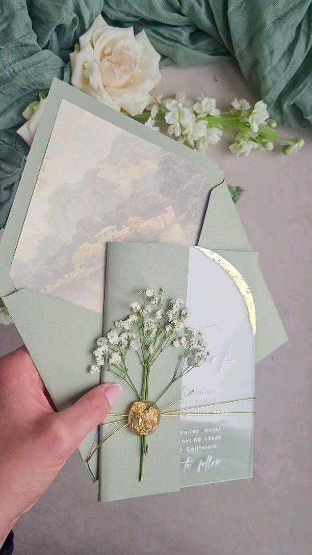 Hand holding a sage green wedding invitation decorated with dried baby's breath and gold accents.