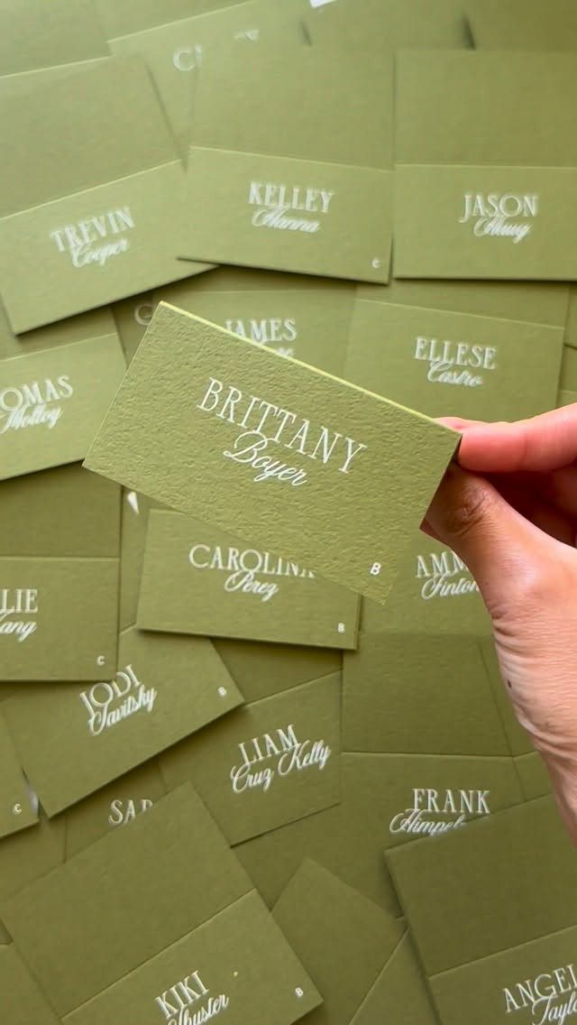 A hand holds a sage green textured place card with white calligraphy names.