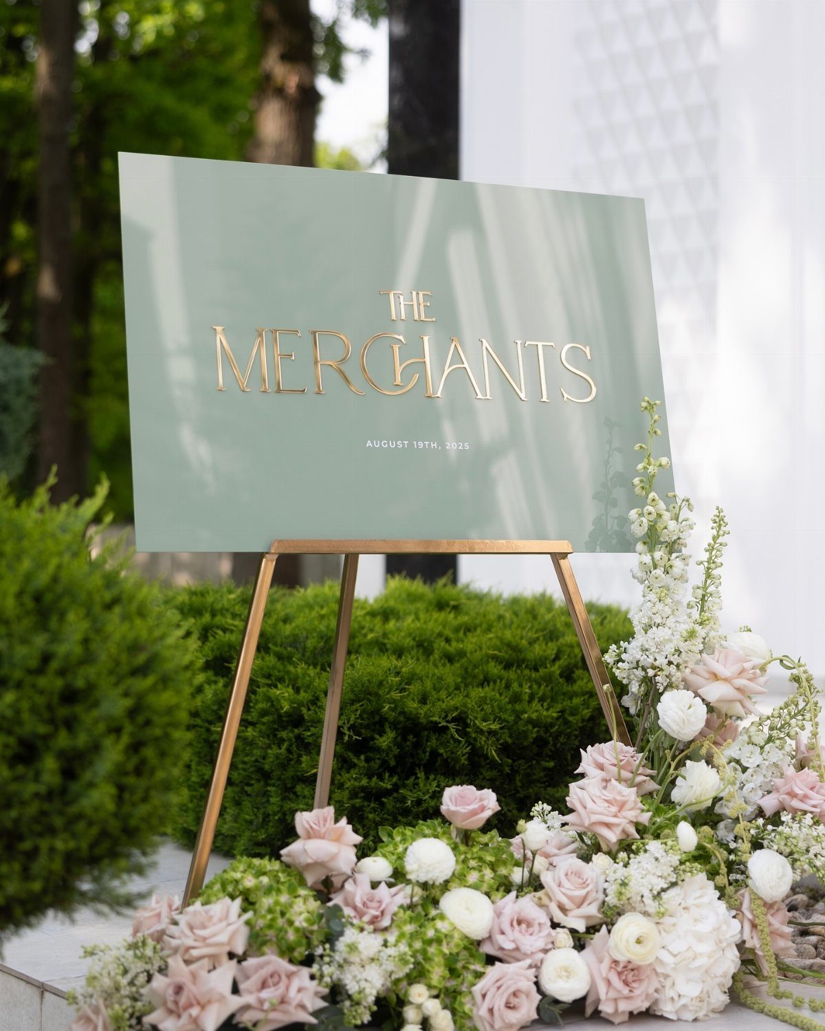 Sage green wedding sign with gold text displayed on a gold easel among flowers.