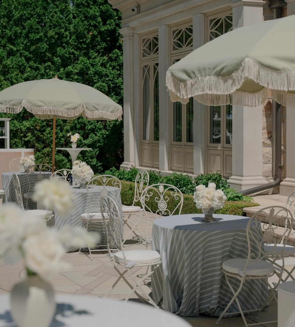 Sage green fringed patio umbrellas shade outdoor tables with white floral arrangements.