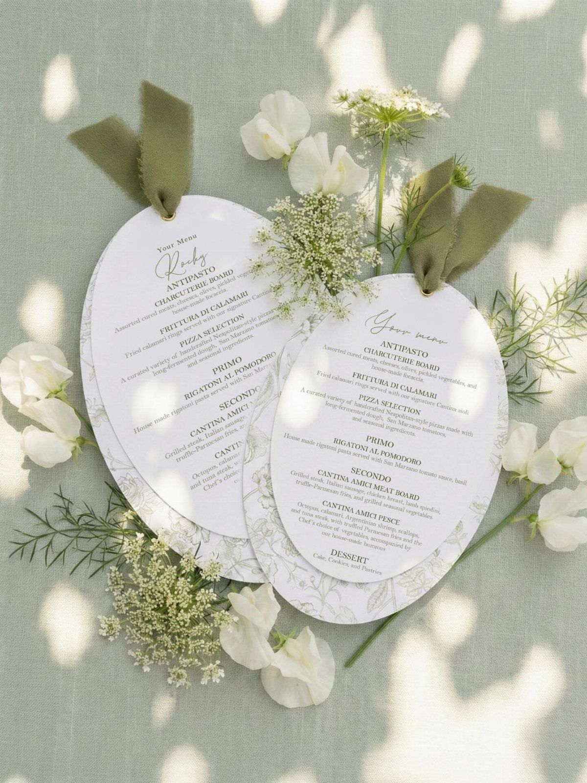 Two white oval wedding menus decorated with white flowers and sage green ribbons on linen.