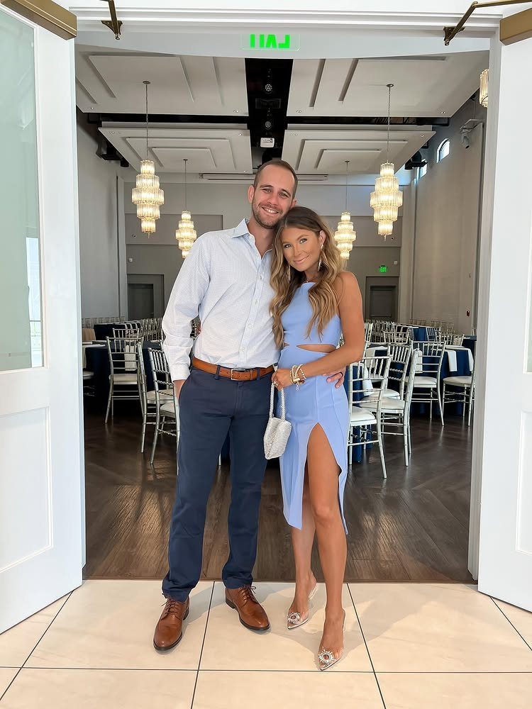 A smiling man in a button-down shirt and navy pants stands with a woman in a blue dress.
