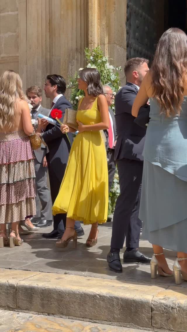 People dressed in semi-formal wedding attire standing outside a stone building in bright daylight.