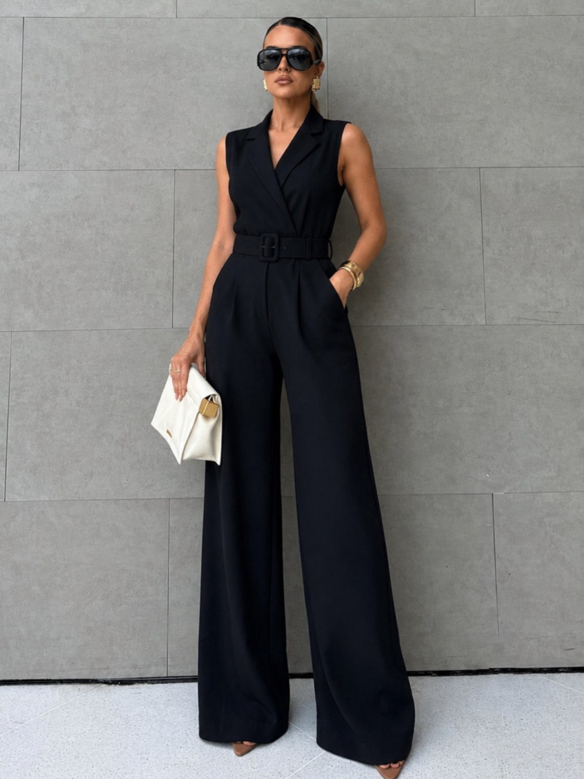 A woman in a black sleeveless wide-leg jumpsuit posing against a grey wall.