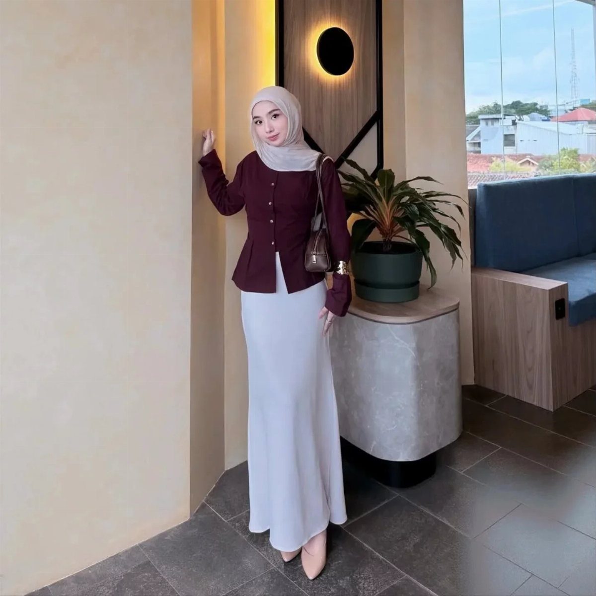 Woman in a maroon blouse and long white skirt posing indoors near a plant.