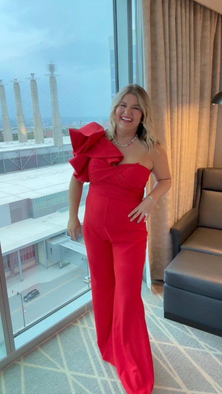 A smiling woman posing in a vibrant red one-shoulder jumpsuit with a large ruffle detail.