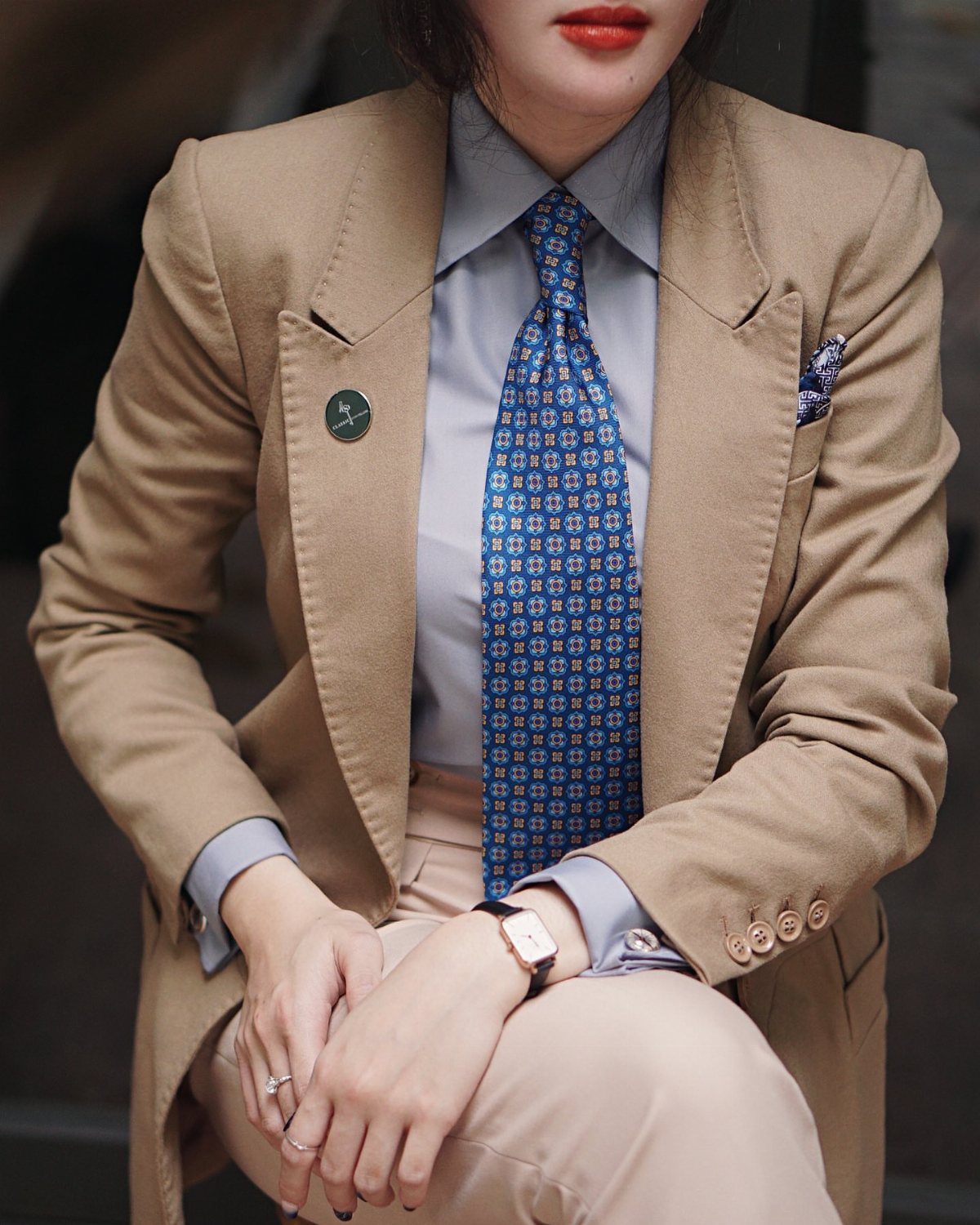 A person wearing a tan blazer, blue patterned tie, and matching pocket square with minimal jewelry.