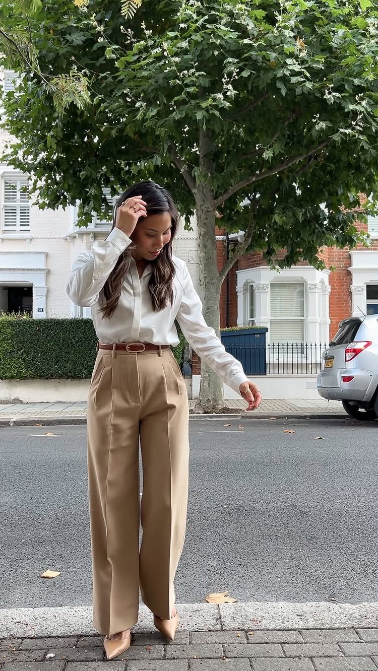 A woman posing in a white shirt, tan wide-leg trousers, and tan heels on a street.