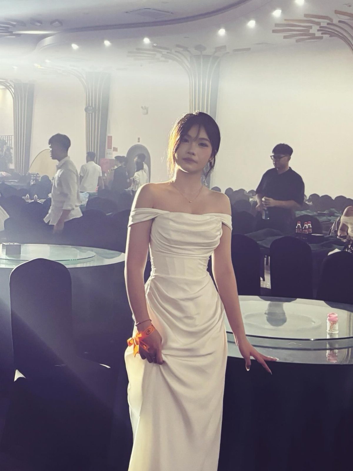 A woman wearing a white off-the-shoulder satin gown standing in an event hall.