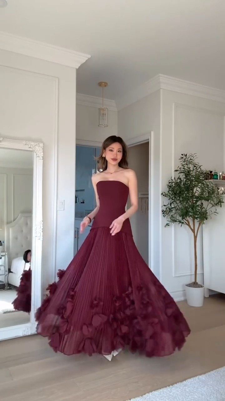 A woman posing in a voluminous, strapless burgundy ballgown with floral details on the skirt.