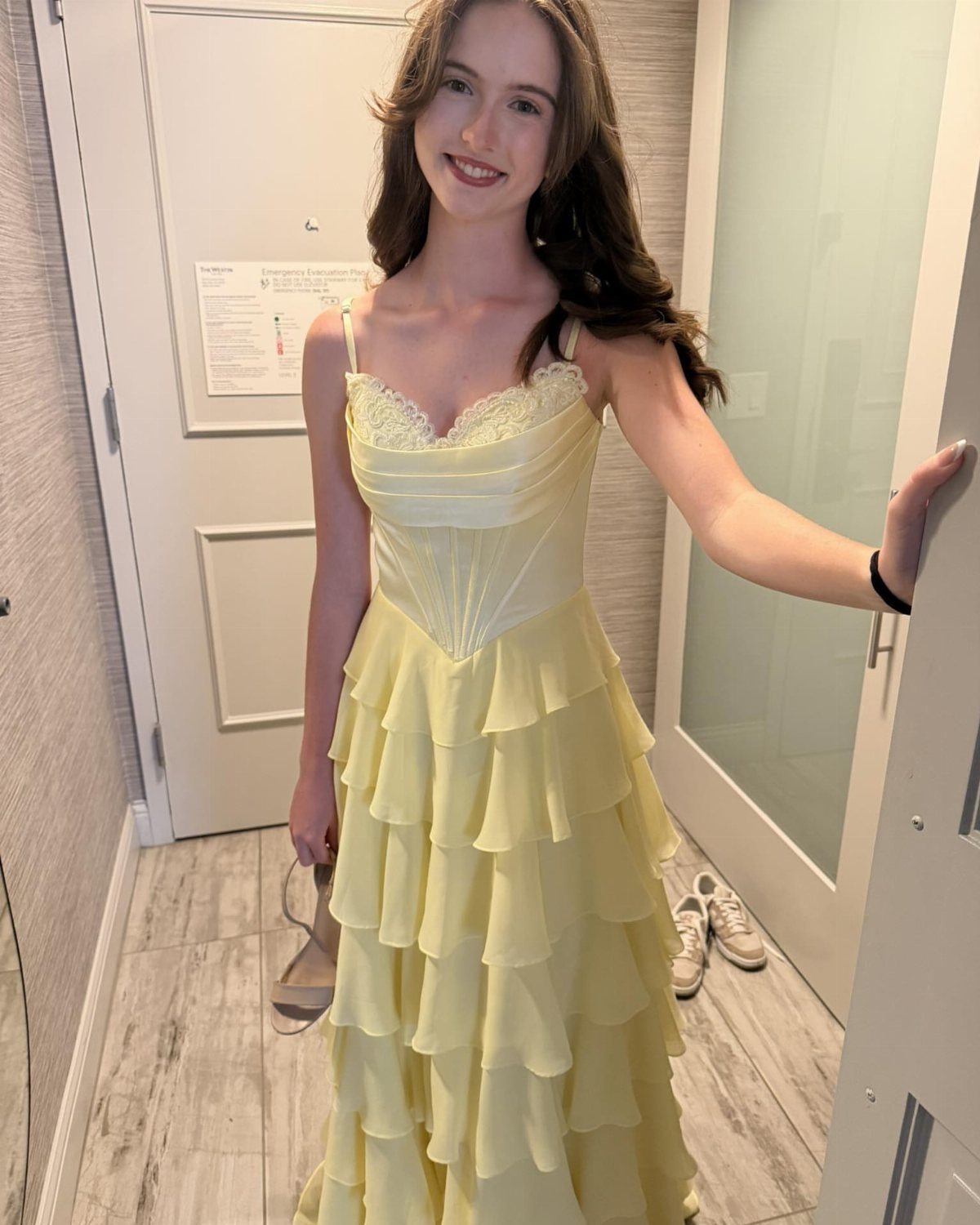 A young woman smiling in a yellow tiered ruffled prom dress standing in a room.