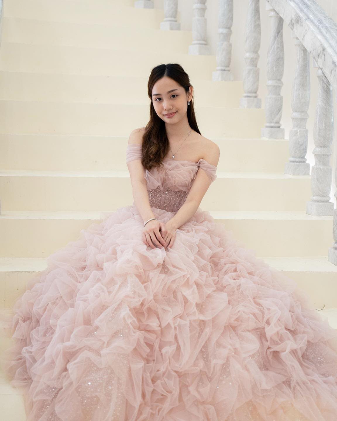 A young woman in a blush pink ruffled tulle ballgown sitting on white stairs.
