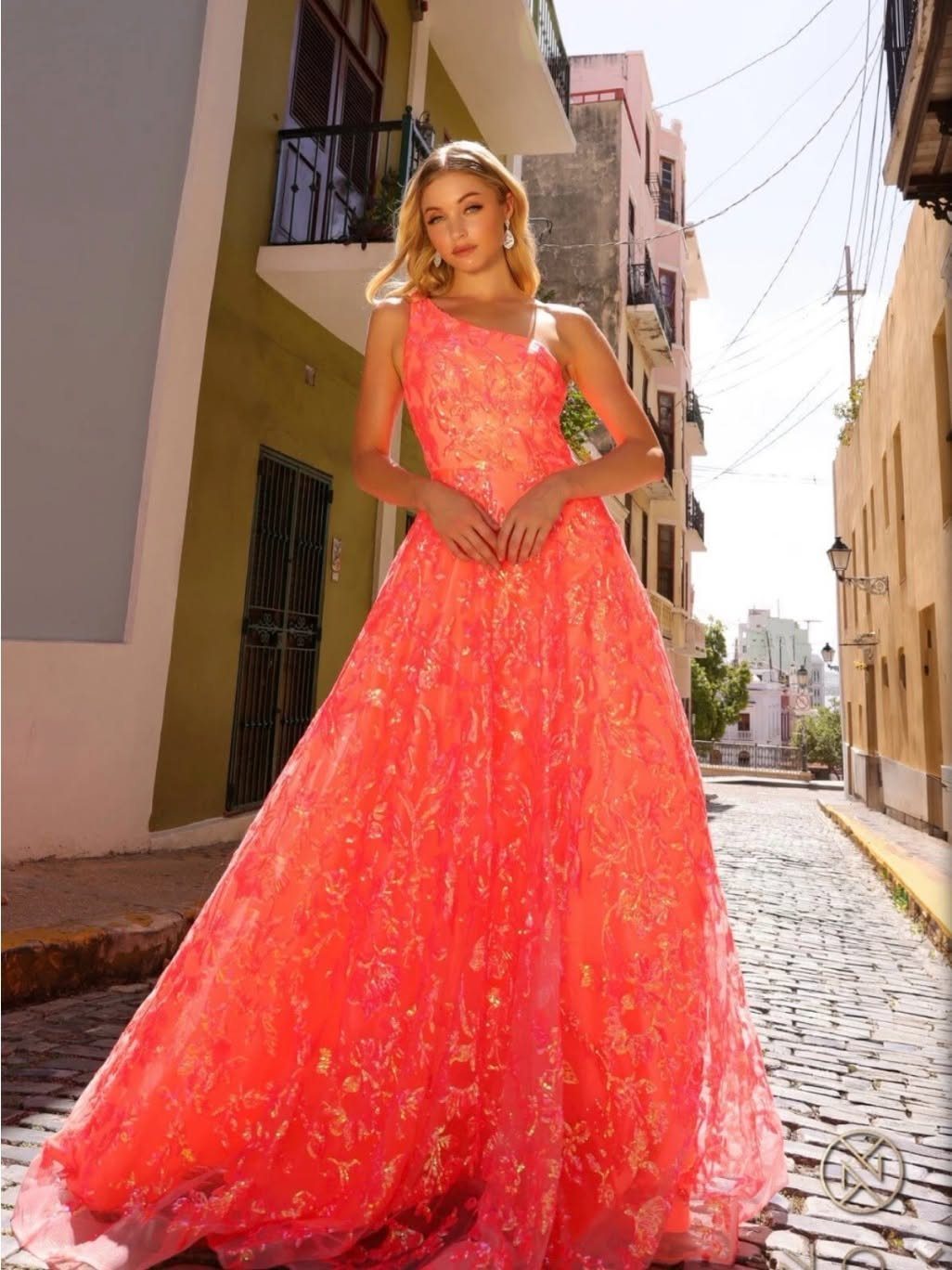 Young woman wearing a vibrant coral pink embroidered one-shoulder prom dress on a cobblestone street.