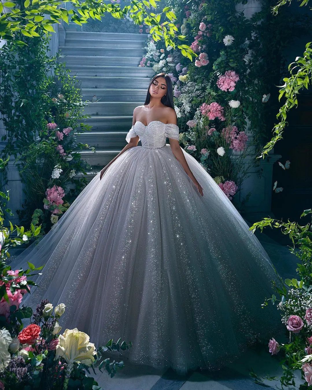 Woman in a shimmering white ball gown wedding dress posing amidst flowers and stone stairs.