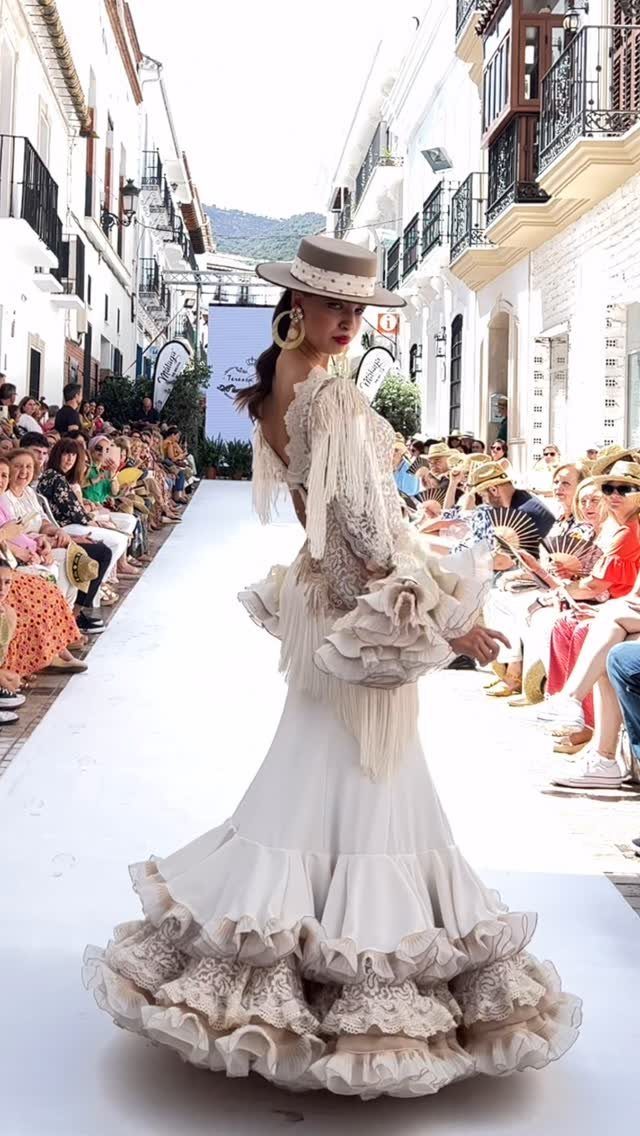 Model walking a runway in a white ruffled wedding dress with fringe and a wide-brimmed hat.