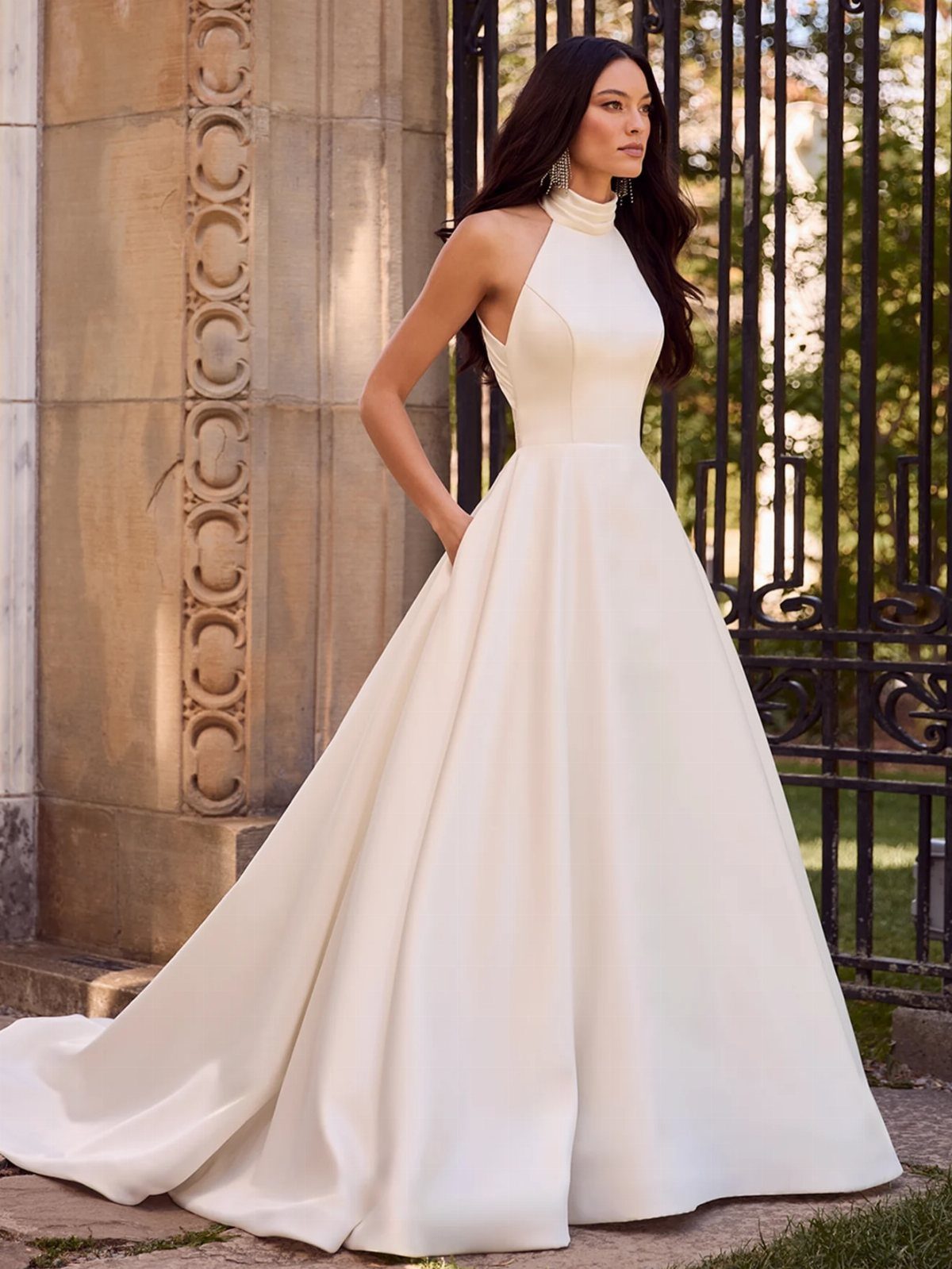 Woman in a white sleeveless satin wedding gown standing by a stone pillar and iron gate.