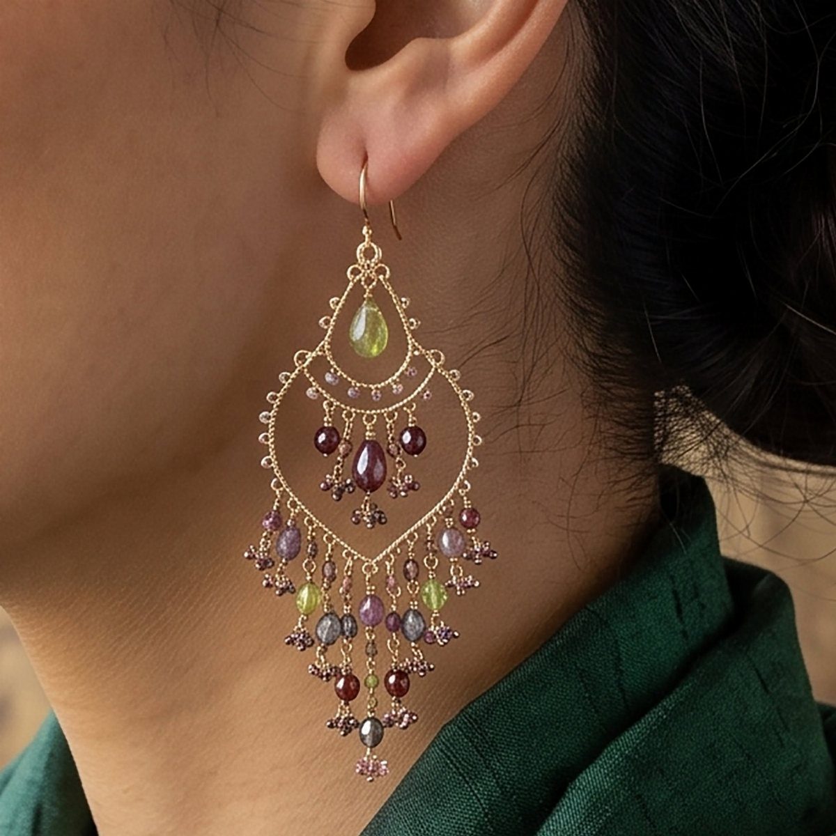 Large gold chandelier earrings with green and purple gemstones hanging from a person's ear.