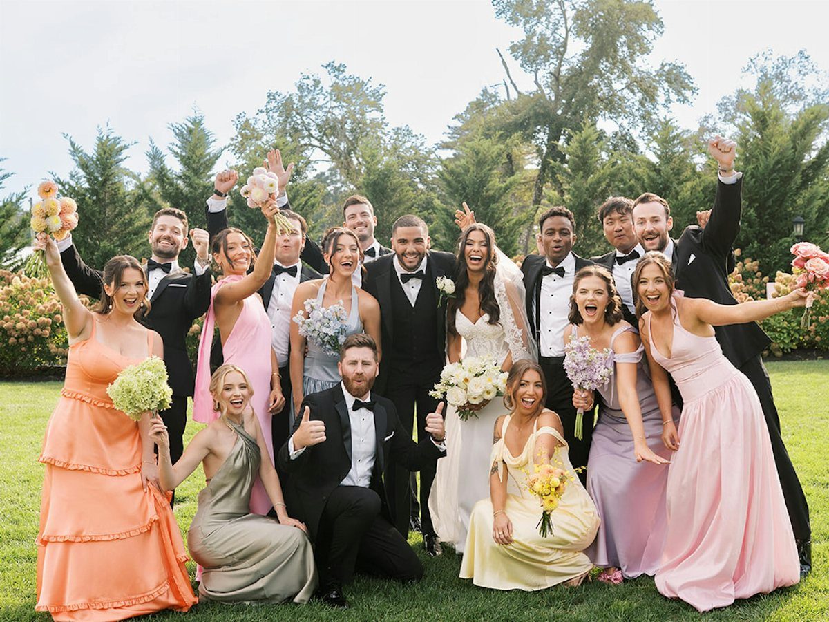 A wedding party posing in a field wearing colorful pastel bridesmaid dresses and black tuxedos.