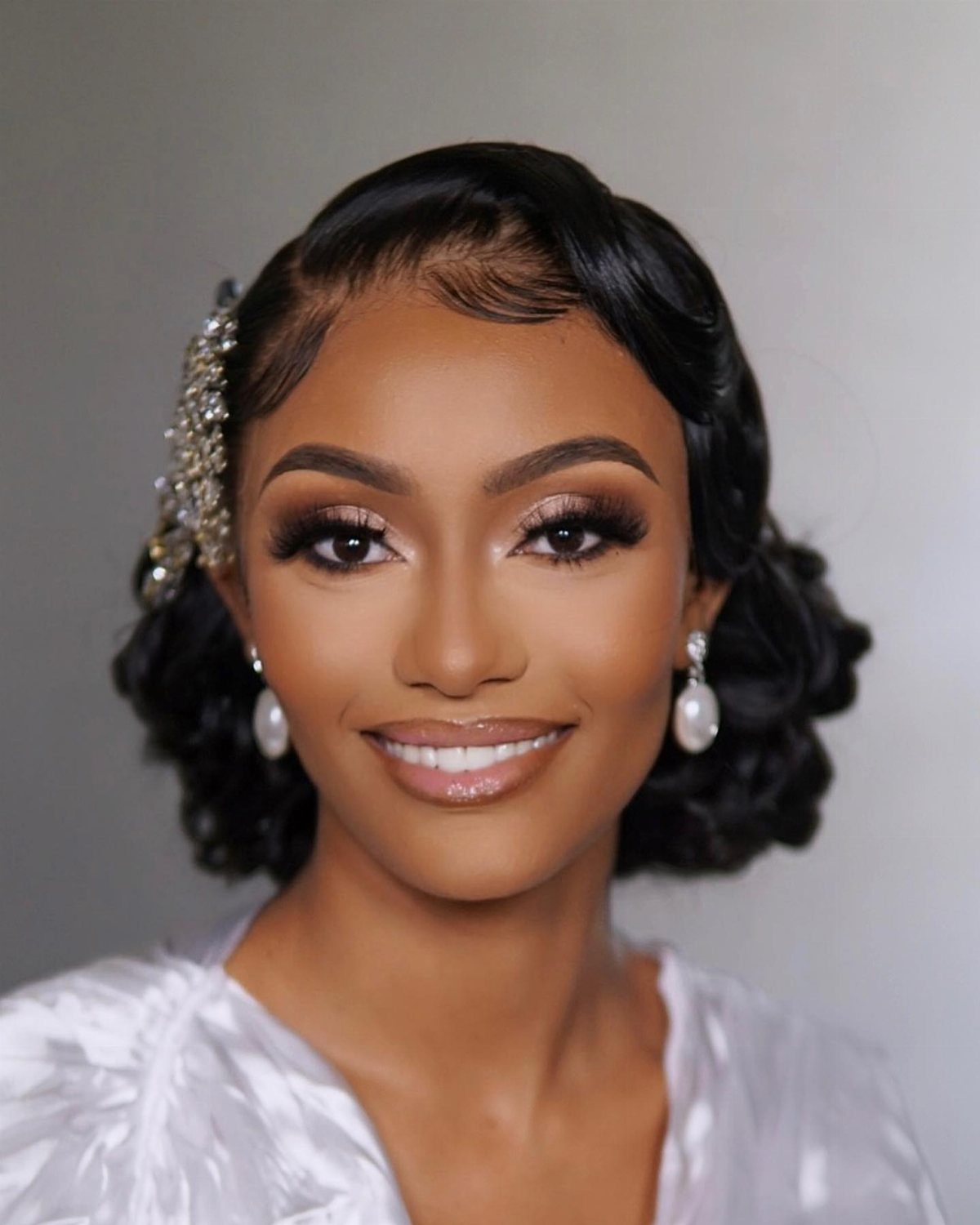 A smiling woman with vintage wavy hair, pearl earrings, and glamorous makeup looking at the camera.
