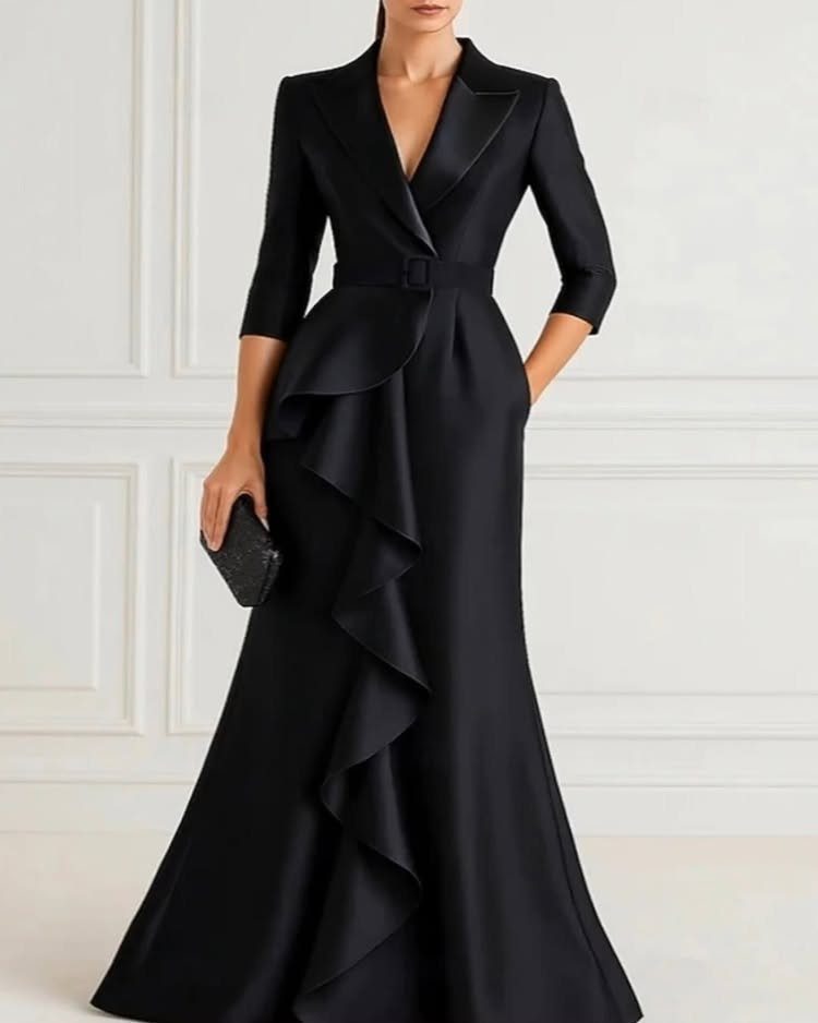 Woman in a black satin gown with large side ruffles and a belted waist standing against a wall.
