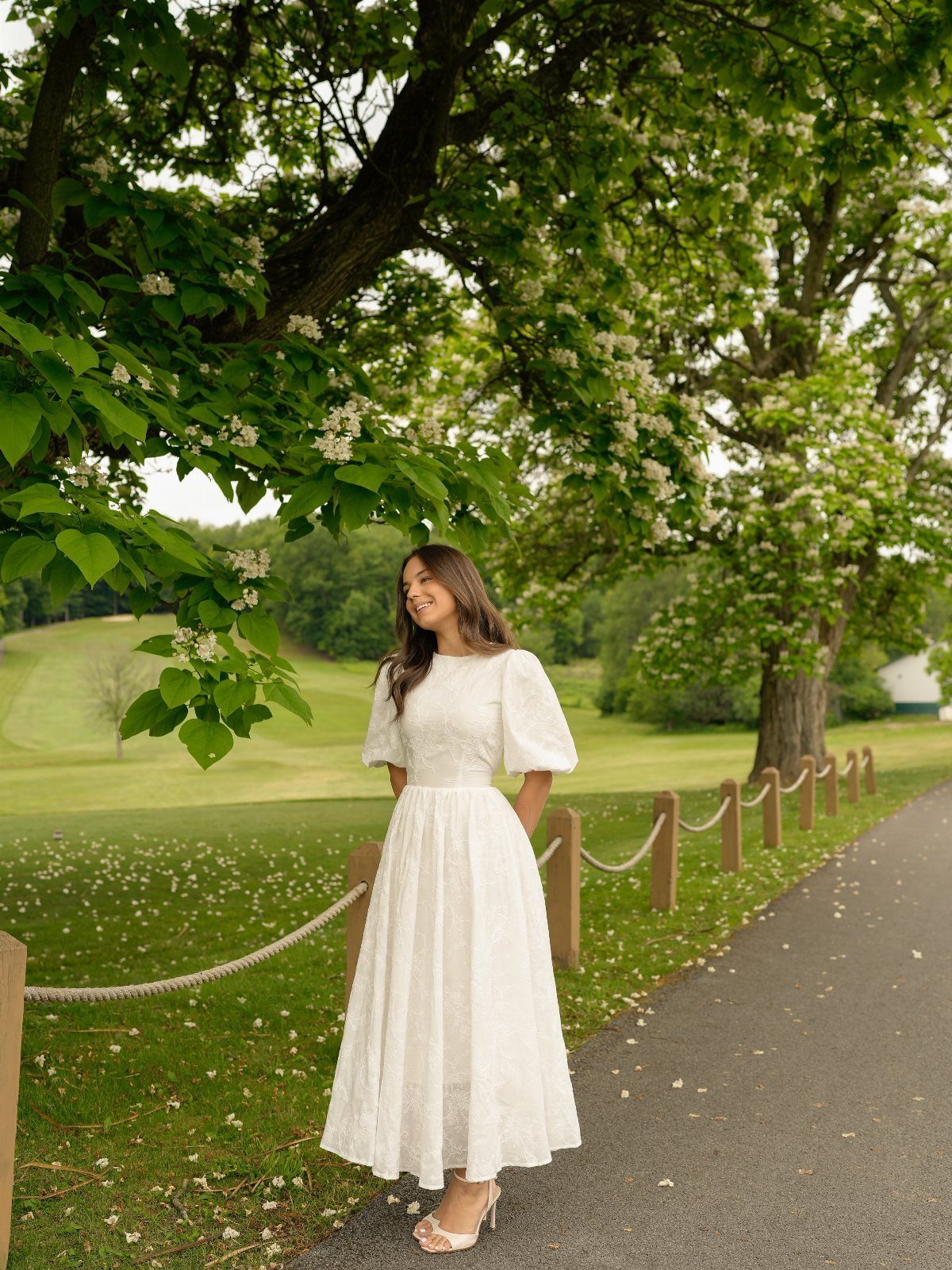 A woman in a white puff-sleeve midi dress standing on a path under a tree.