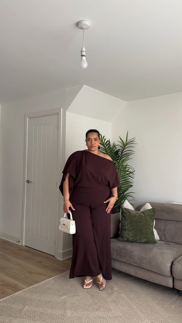 Woman posing in a dark burgundy one-shoulder jumpsuit and white sandals.