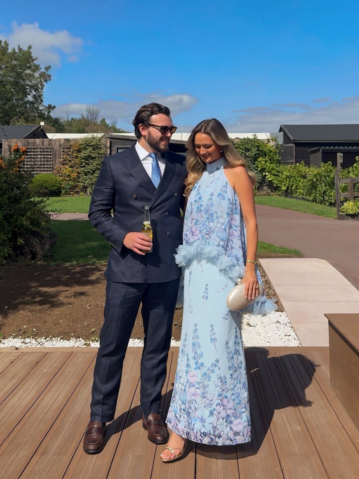 A man in a navy suit and a woman in a light blue floral maxi dress standing outdoors.