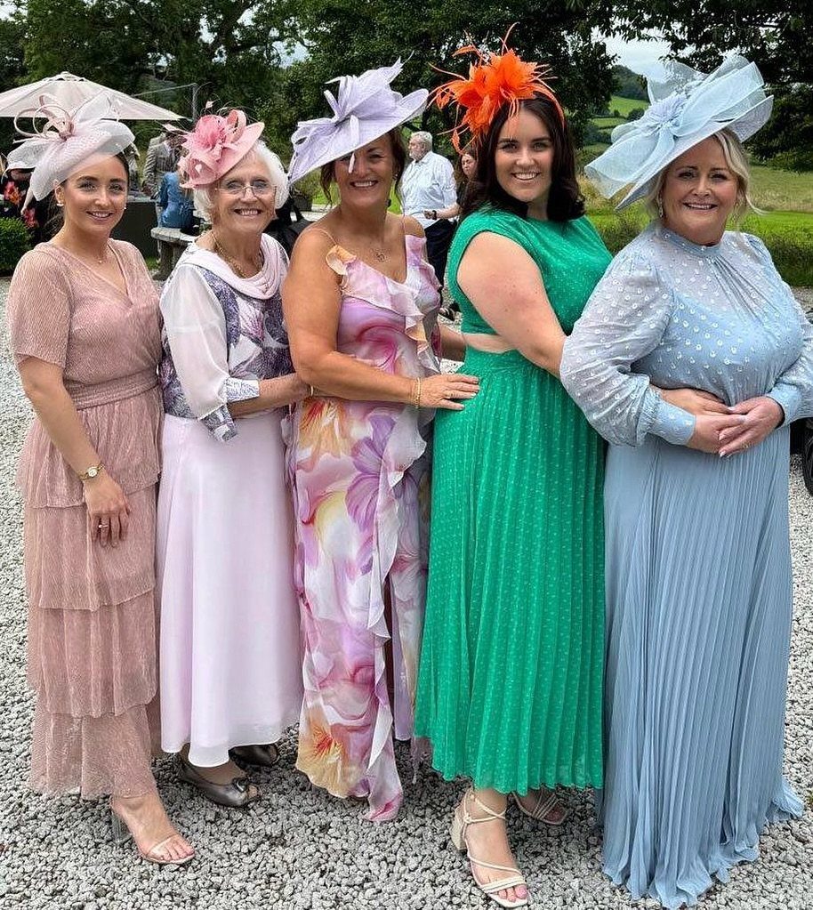 Five women in colorful spring dresses and large fascinators standing on a gravel path outdoors.