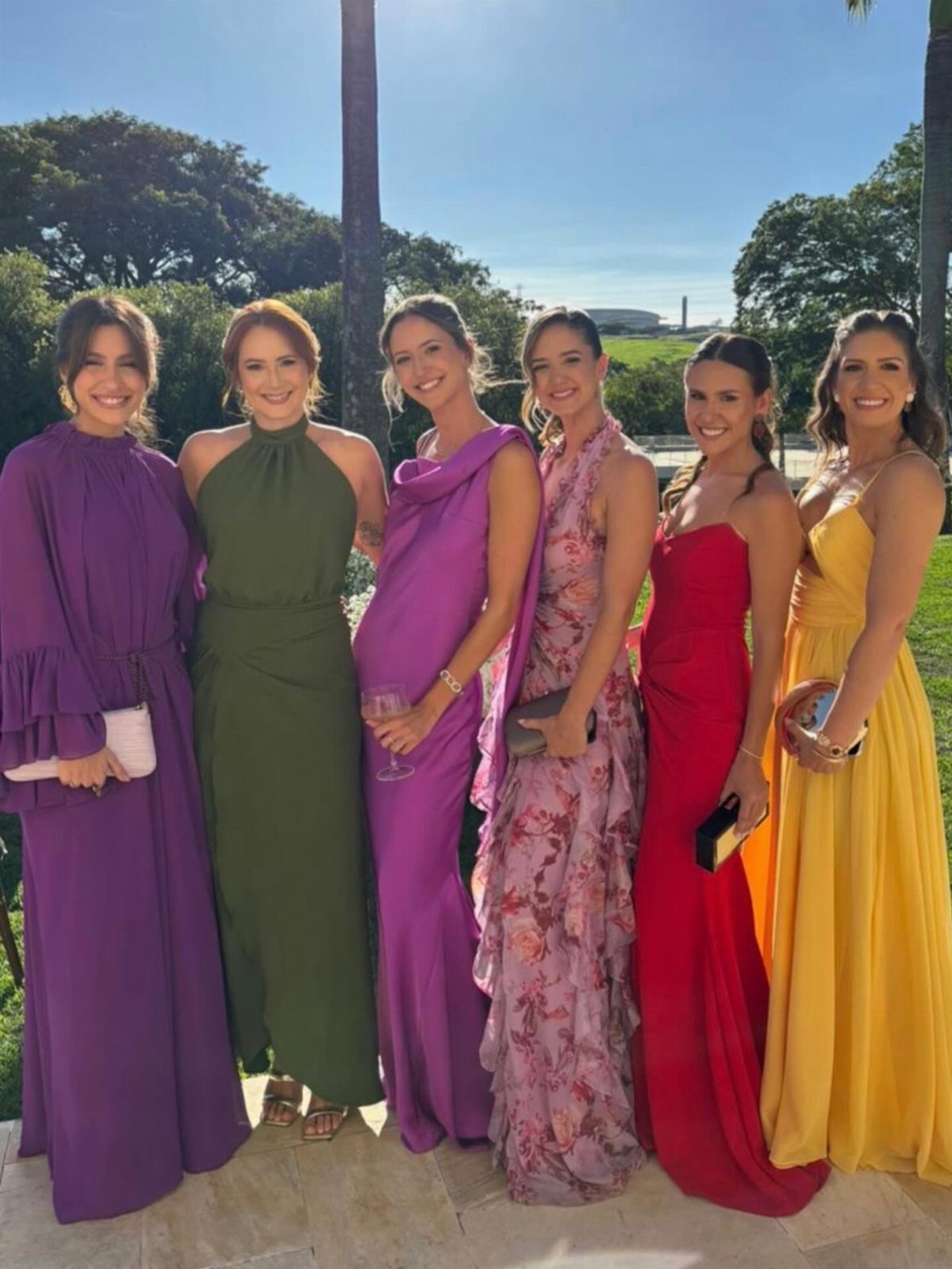 Six women posing together in vibrant, colorful long formal dresses in an outdoor garden setting.