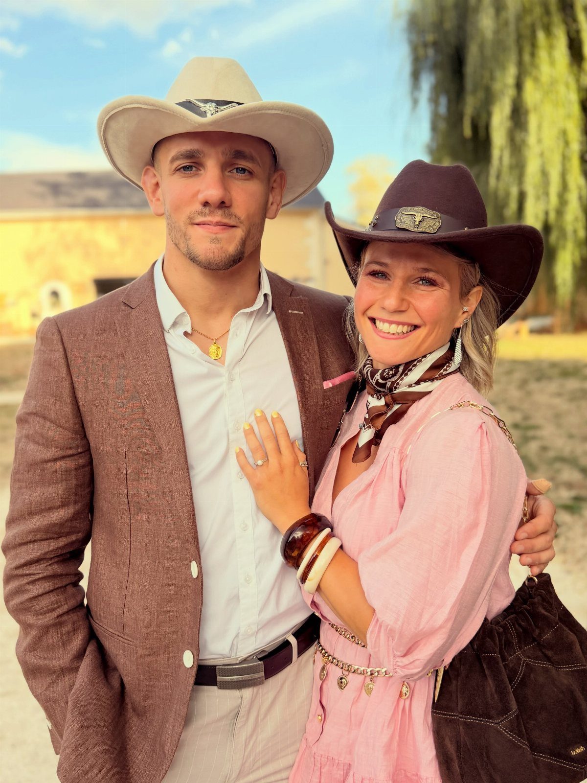 A smiling man and woman wearing cowboy hats and stylish western-inspired attire outdoors.