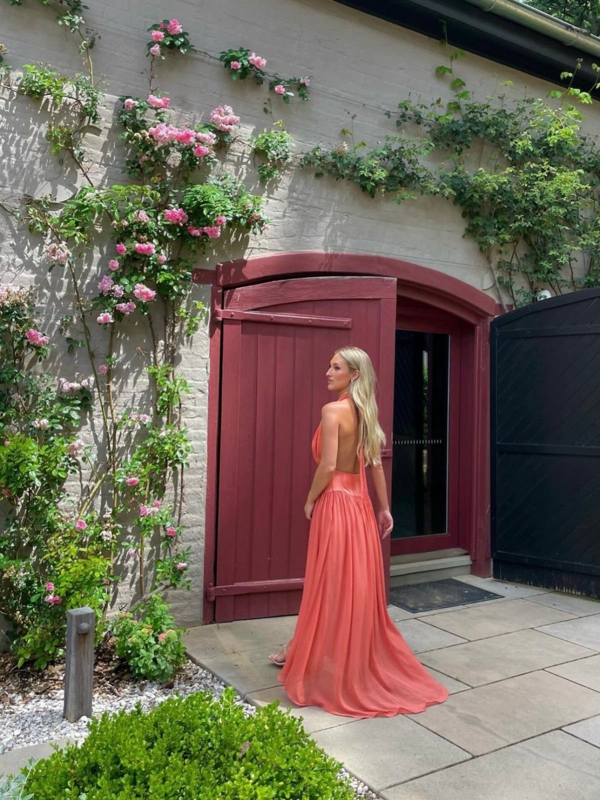Blonde woman wearing a long coral backless dress standing by a red door and rose vines.
