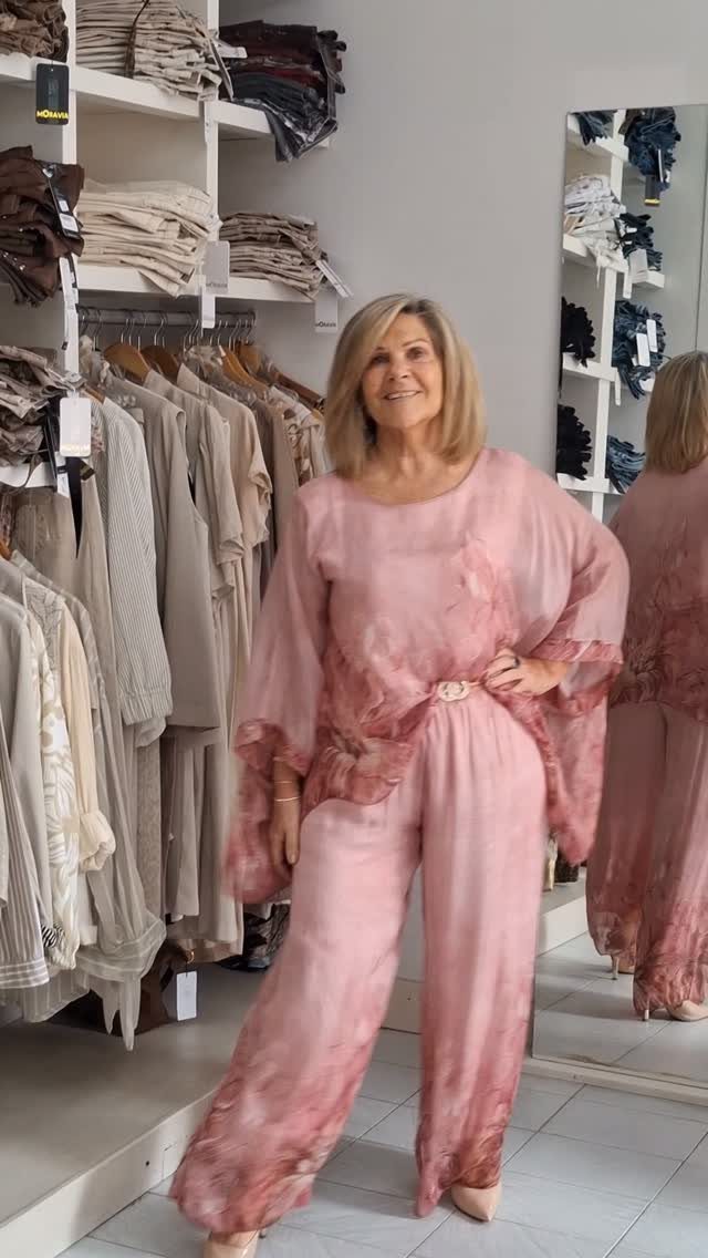 Smiling older woman posing in a pink flowing two-piece outfit inside a clothing boutique.