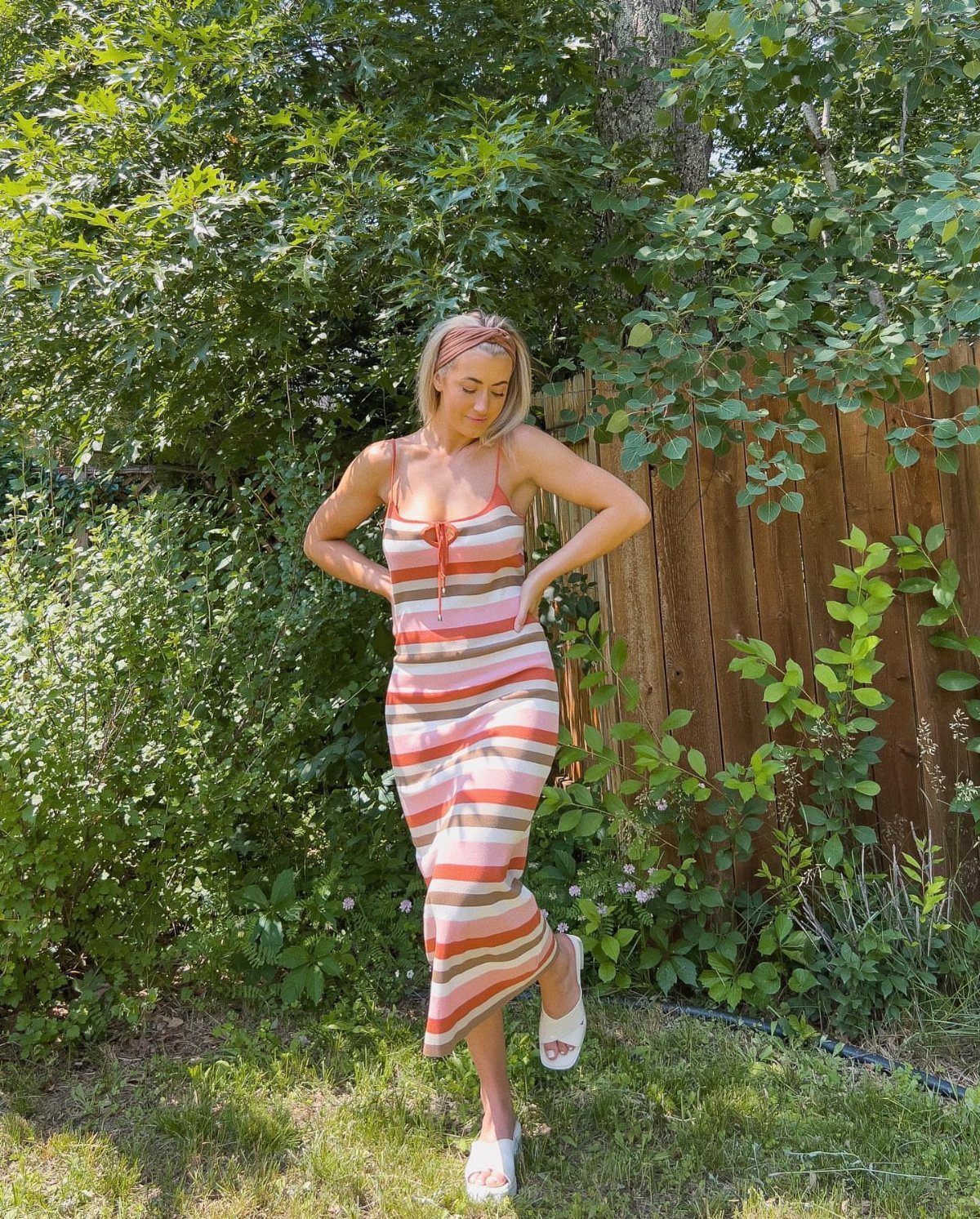 Woman posing in a garden wearing a long, striped summer dress with orange and white patterns.