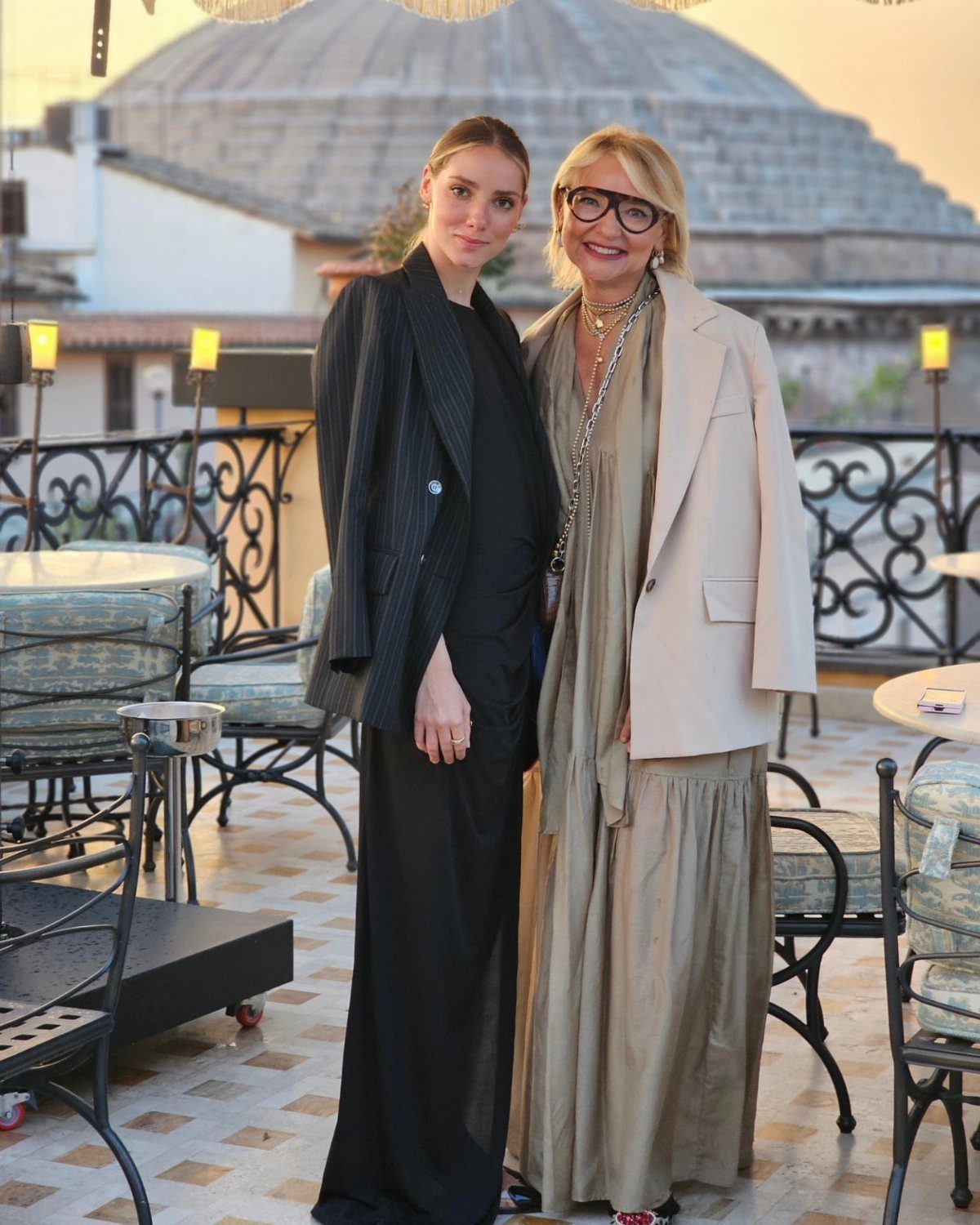 Two women posing on a terrace wearing elegant dresses and tailored blazers at sunset.